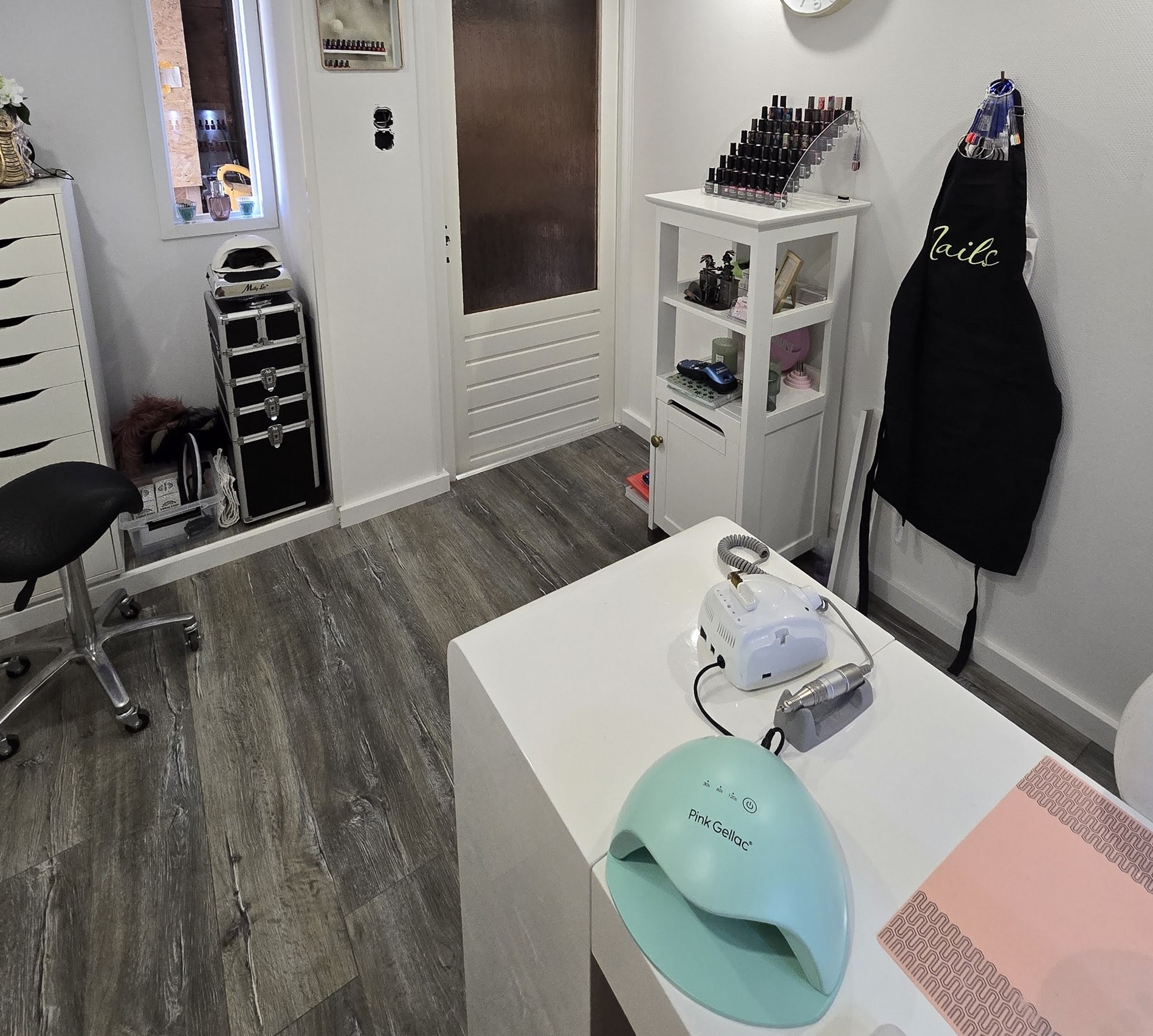 De salon: de behandeltafel met nagellamp, een rek vol nagellak en een zwart schort met Nails erop