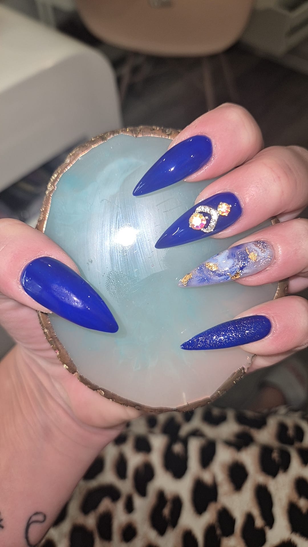 Kobaltblauwe stiletto nagels met een gouden bedel, vastgehouden boven een blauwe agaatschaal